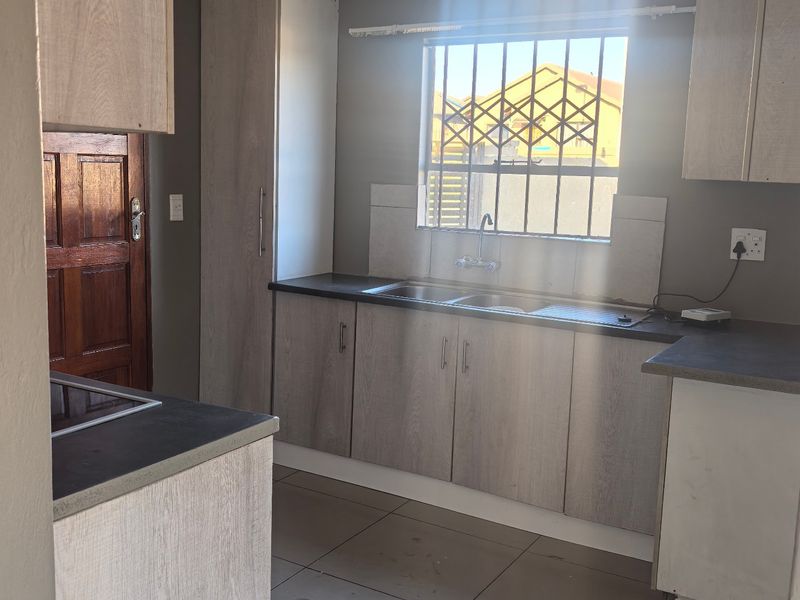 A functional kitchen featuring light wood-look cabinetry, dark laminate countertops, a stainless steel sink, and tiled flooring with natural light.