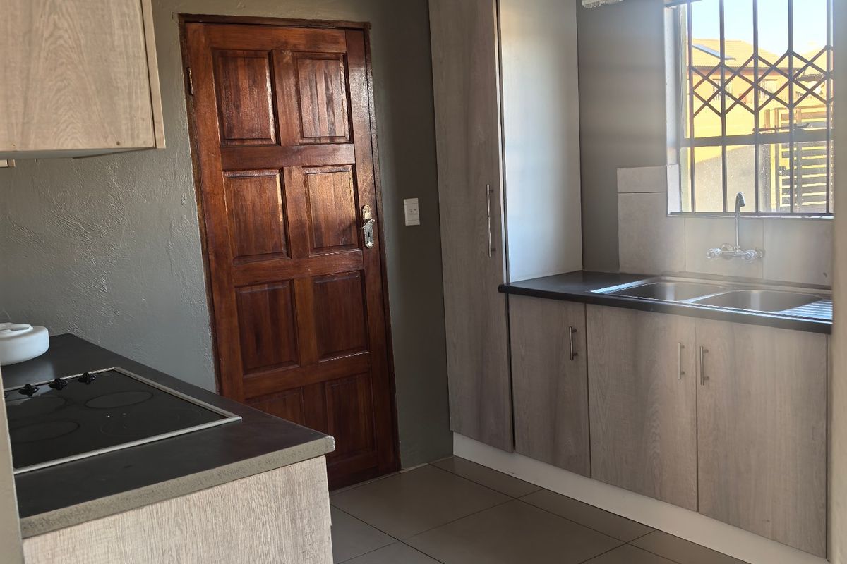 A functional kitchen featuring light wood-look cabinetry, a built-in electric hob, double sink, and tiled flooring with security bars on the window.