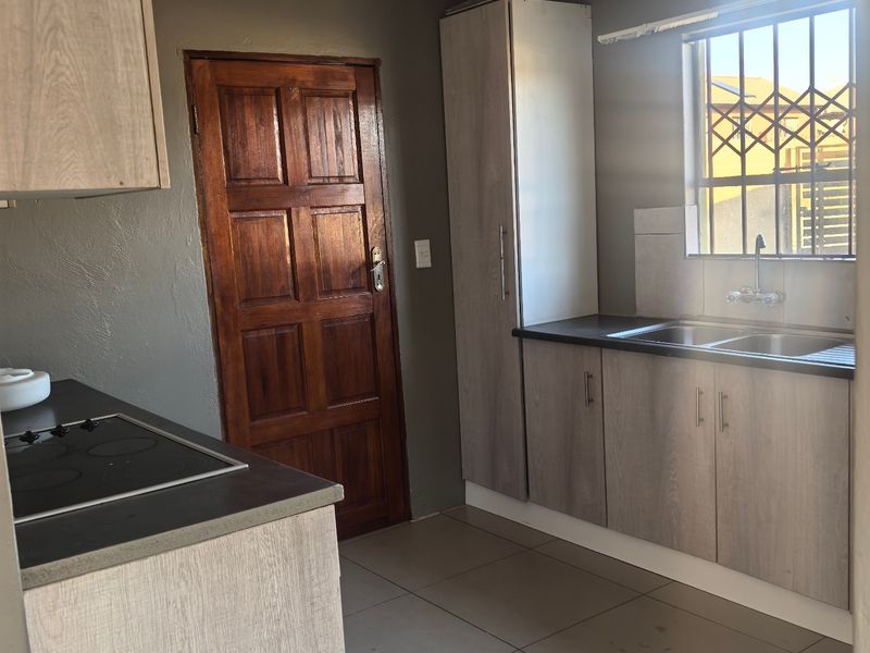 A functional kitchen featuring light wood-look cabinetry, a built-in electric hob, double sink, and tiled flooring with security bars on the window.
