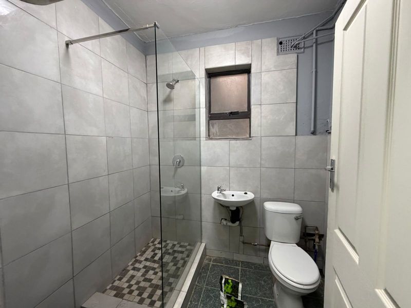 A compact bathroom featuring a glass-enclosed shower, wall-mounted basin, and toilet, with grey tiled finishes and exposed plumbing elements.
