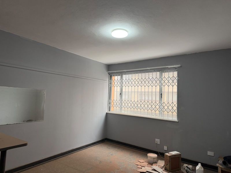 An empty bedroom featuring grey walls, a window with a security gate, and unfinished flooring, currently undergoing renovation.
