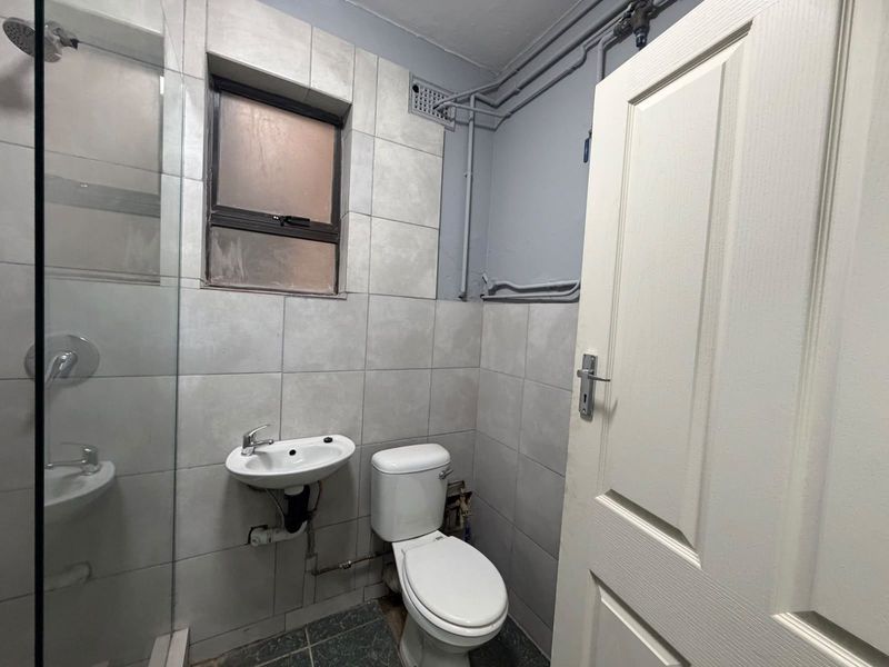 A compact bathroom featuring a shower enclosure, wall-mounted basin, and toilet with exposed surface-mounted plumbing and neutral wall tiling.
