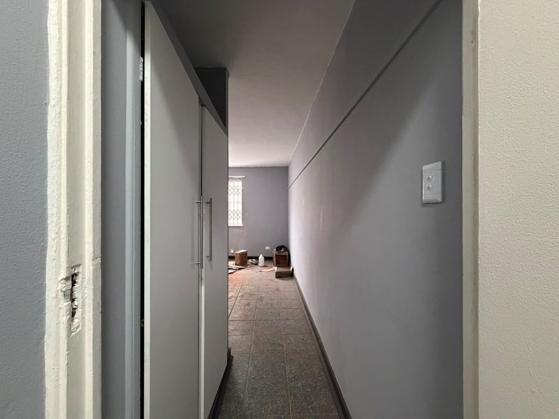 A vacant hallway featuring built-in cupboards, tiled flooring, and a window with security bars, leading into an adjacent room.