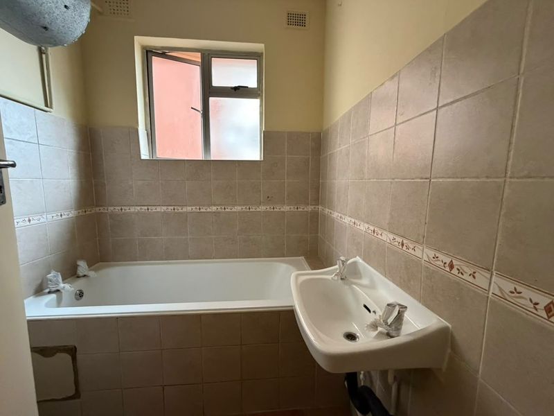 A dated bathroom featuring a standard bathtub, wall-mounted basin, and neutral ceramic wall tiling, requiring cosmetic updates to modernize the space.