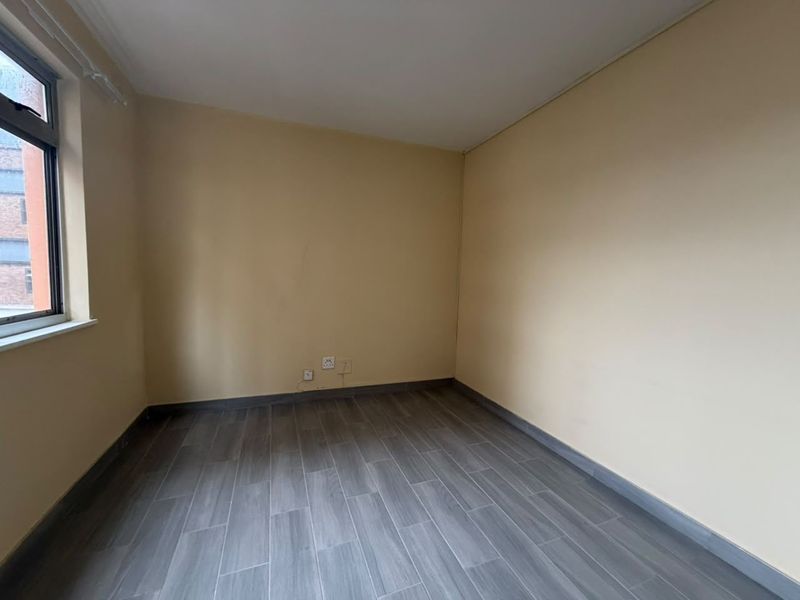 A clean, empty bedroom featuring modern grey wood-look floor tiles, neutral wall paint, and an aluminium-framed window.