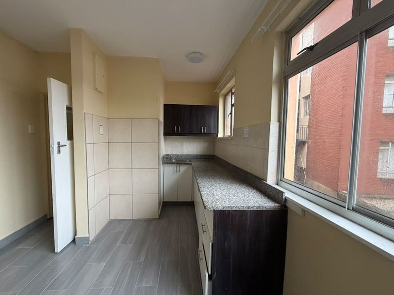 A functional, empty kitchen featuring granite countertops, dark and light cabinetry, tiled splashbacks, and large windows providing natural light.