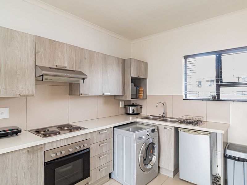 A functional kitchen featuring light wood-grain cabinetry, integrated electric hob and oven, stainless steel sink, and tiled splashback with ample natural light.