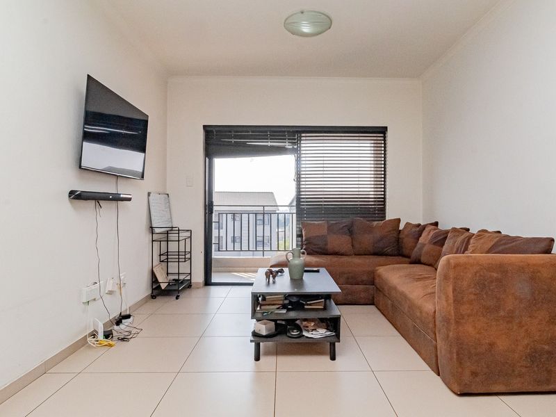 A functional living room featuring neutral tiled flooring, a sliding glass door leading to a balcony, and fitted venetian blinds.