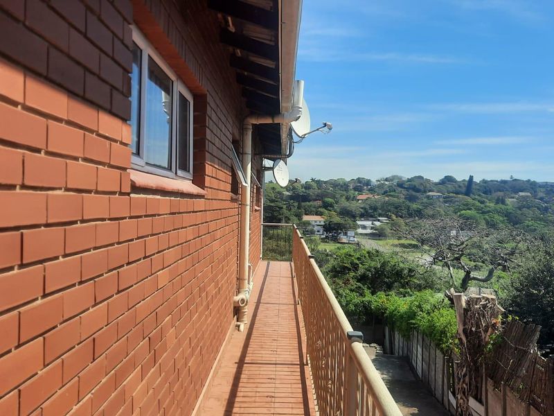 A narrow exterior balcony featuring brick walling, metal railings, and tiled flooring, offering an elevated view of the surrounding neighborhood and greenery.
