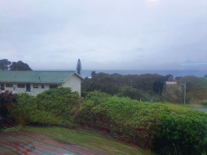 An elevated view overlooking a green metal-roofed building, lush garden foliage, and a distant coastal horizon under an overcast sky.