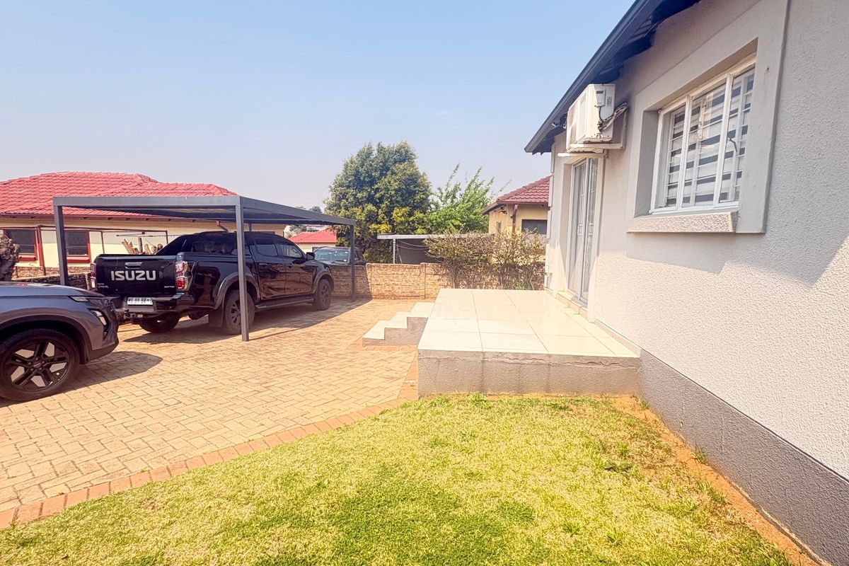 Rear exterior view featuring a paved driveway, carport, and a raised tiled patio area adjacent to the main house structure.