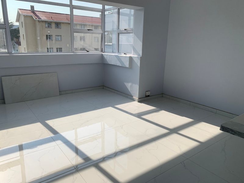 A bright, empty living area featuring large windows and modern light-colored tiled flooring, offering a clean and neutral space ready for furnishing.