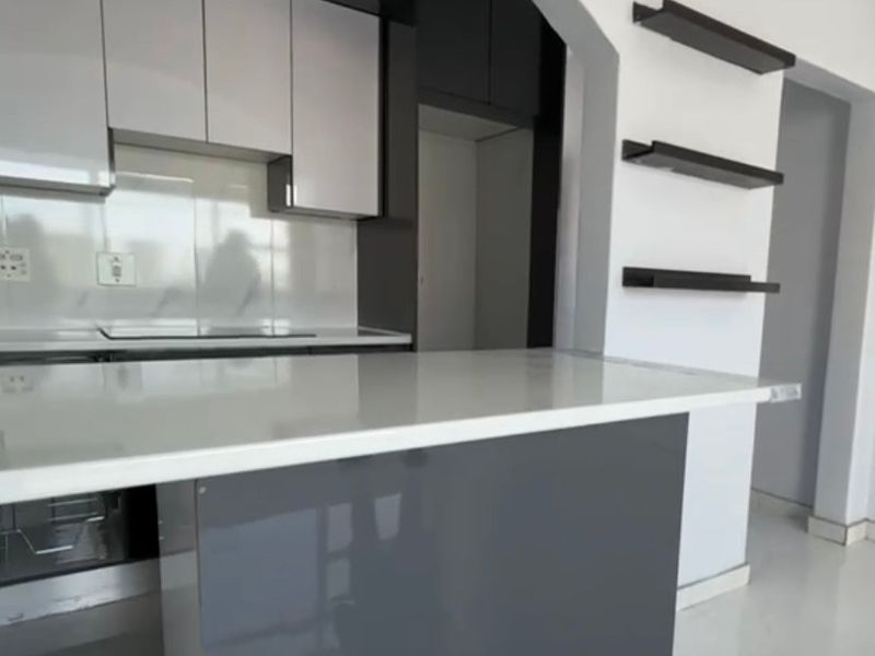 Modern kitchen featuring sleek two-tone cabinetry, stone countertops, a breakfast bar, and decorative wall shelving framed by an elegant architectural archway.