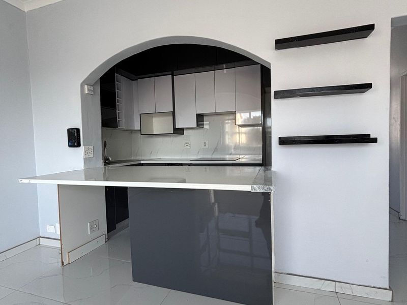 A compact kitchen featuring modern two-tone cabinetry, a stone breakfast bar, and an arched opening, finished with polished floor tiles.