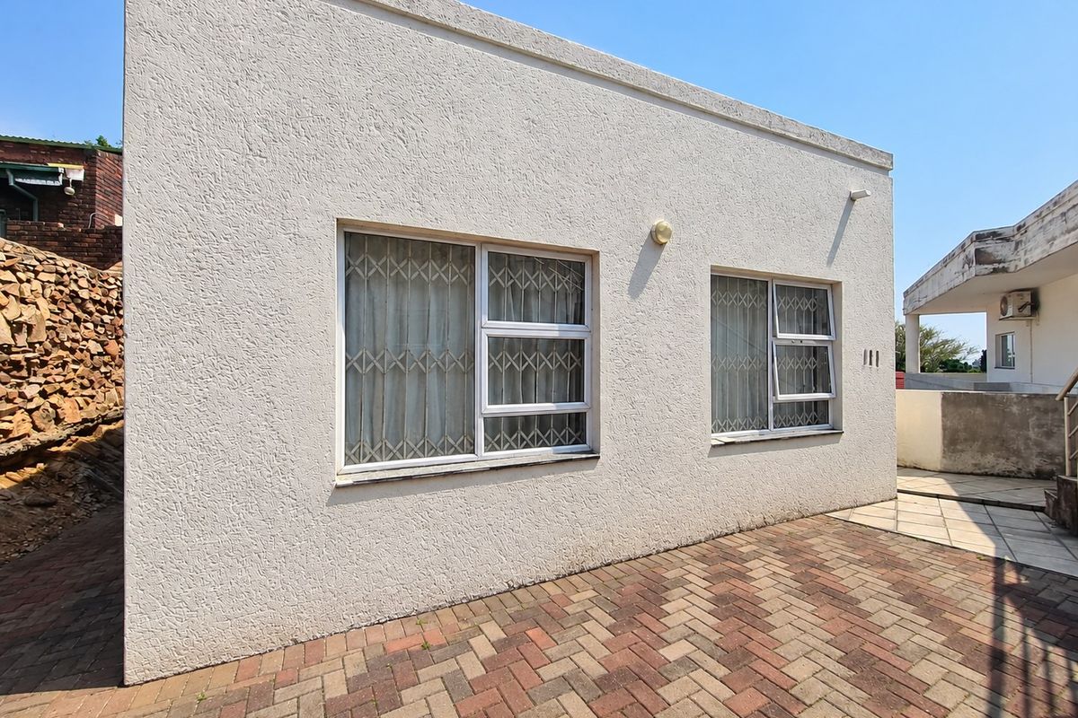 Rear exterior view of the property featuring a textured stucco facade, security-fitted windows, and a paved patio area with a stone retaining wall.