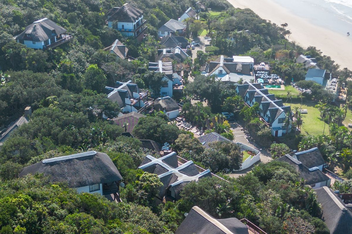An aerial view of a coastal estate featuring multiple residential units with traditional thatched roofs nestled within lush, mature vegetation near the beach.