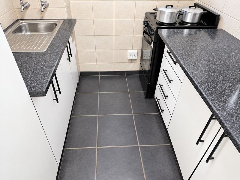 A compact, functional kitchen featuring white cabinetry, dark laminate countertops, a stainless steel sink, and a freestanding stove with tiled splashbacks.