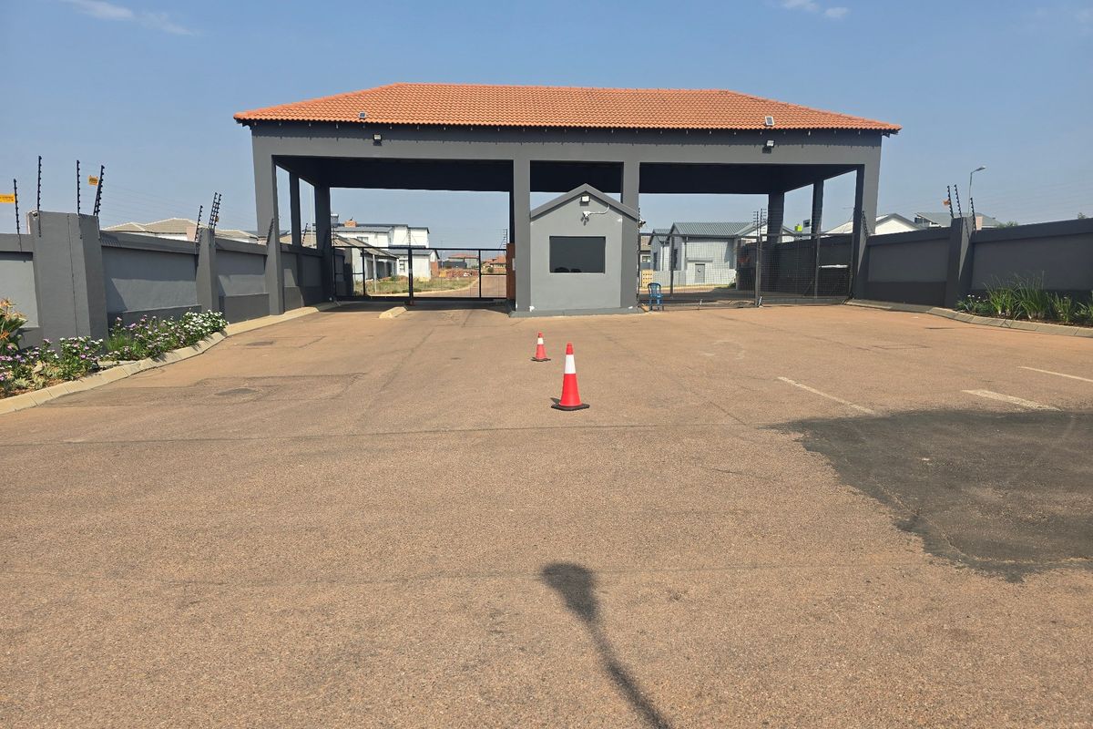 A secure residential estate entrance featuring a gated guardhouse, perimeter walling with electric fencing, and a paved driveway approach.