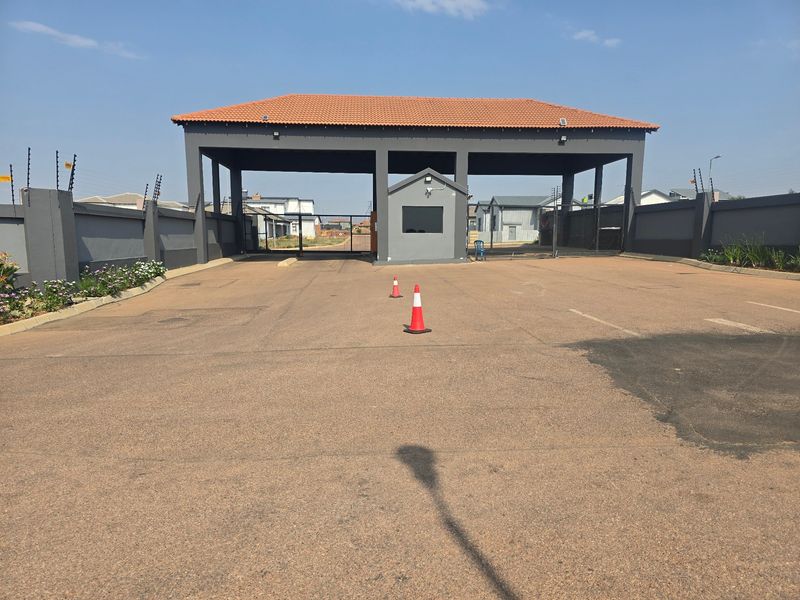 A secure residential estate entrance featuring a gated guardhouse, perimeter walling with electric fencing, and a paved driveway approach.