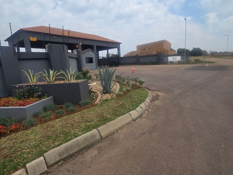 View of the secure estate entrance featuring a modern gatehouse, electric fencing, paved driveway, and well-maintained landscaped garden beds.