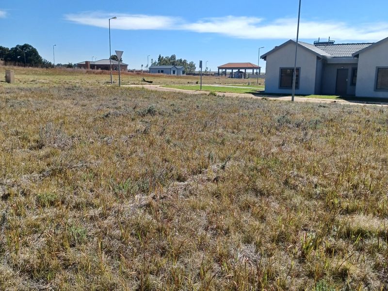 A view of a vacant residential plot with natural grass cover, adjacent to existing residential infrastructure and street lighting.