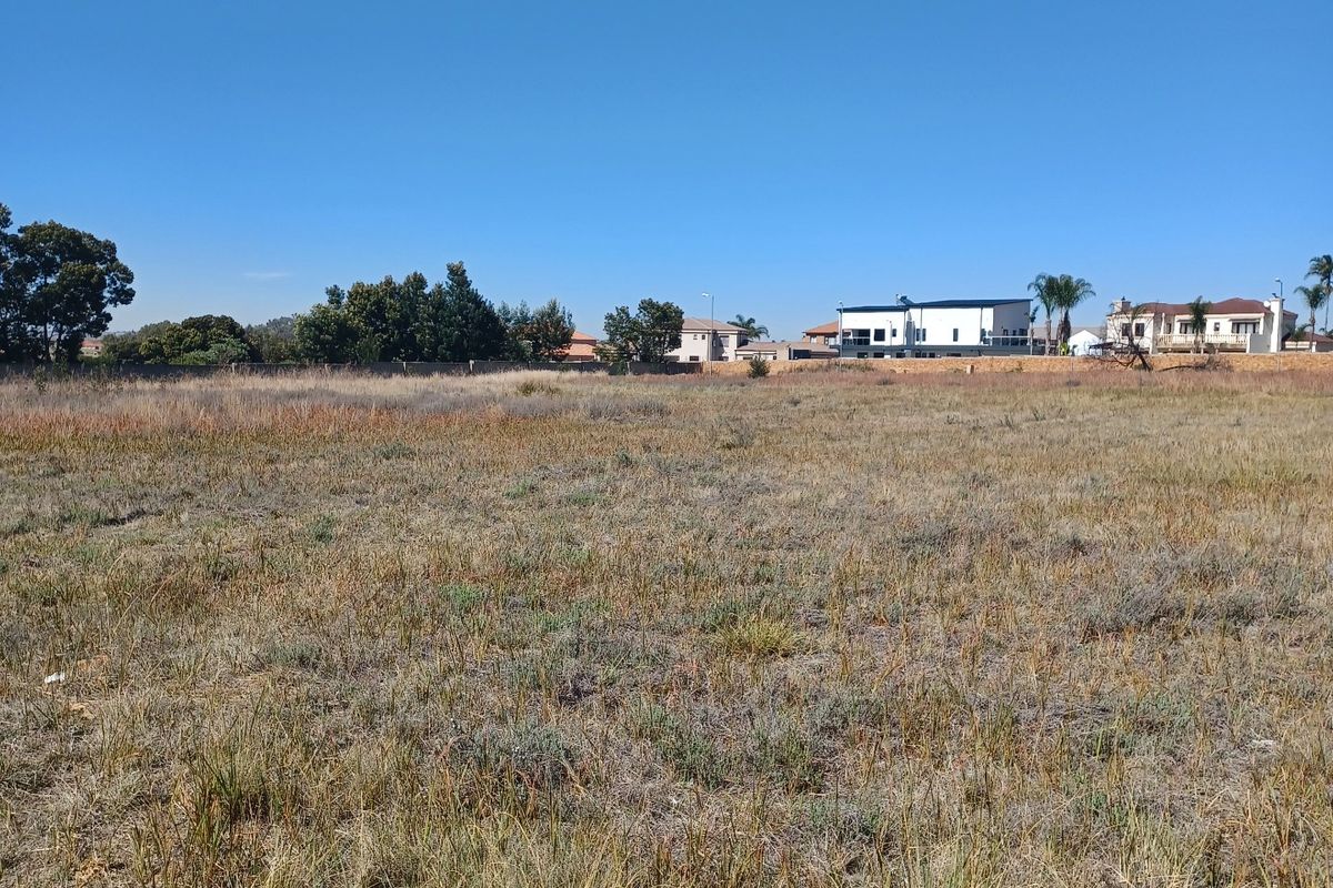 A wide-angle view of a vacant residential plot featuring dry grass and a perimeter boundary wall in the background.