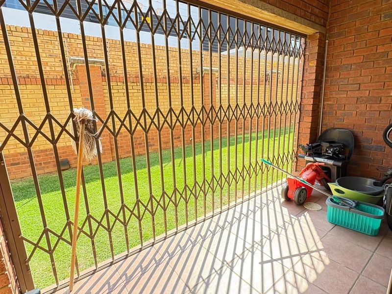 A covered patio area featuring brick walls, tiled flooring, and a full-height security gate overlooking a small garden space.
