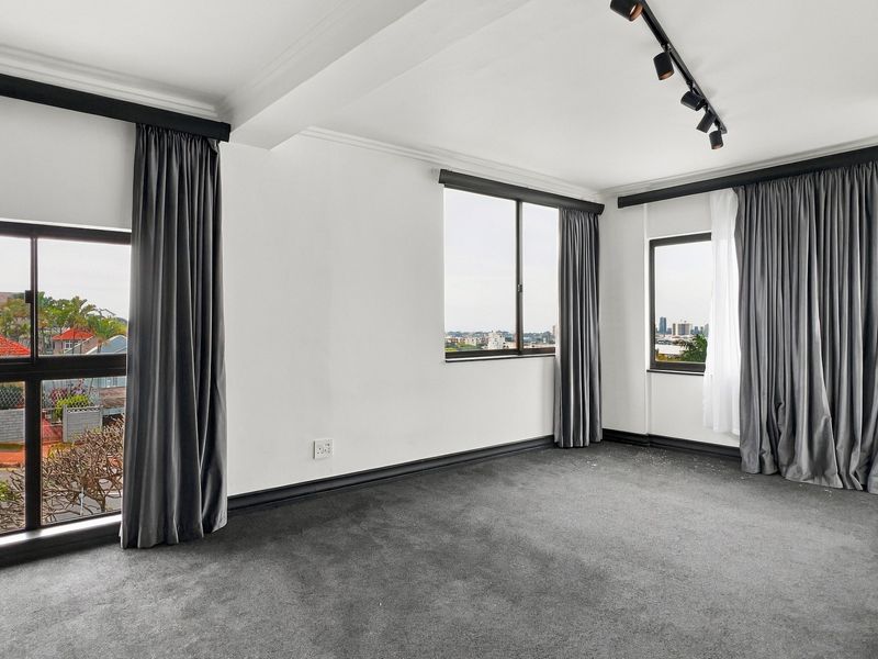 A bright, empty bedroom featuring grey carpeting, track lighting, and built-in cupboards with large windows offering urban views.