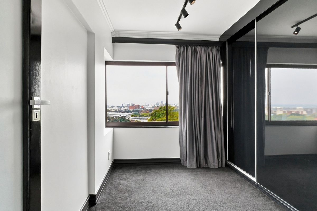 A modern, empty bedroom featuring dark carpet, built-in sliding door wardrobes, track lighting, and a window with city views.
