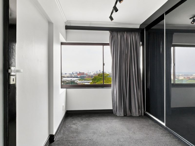 A modern, empty bedroom featuring dark carpet, built-in sliding door wardrobes, track lighting, and a window with city views.