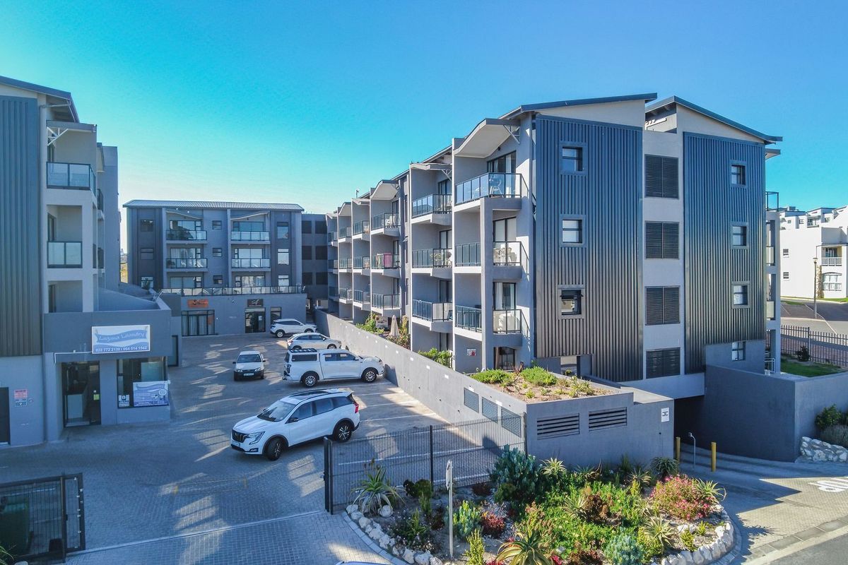 Aerial view of a modern apartment complex featuring metal cladding, private balconies, and a paved parking area with security access.