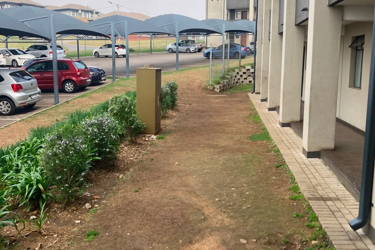 Exterior view of the apartment building showing a paved walkway, adjacent parking area with shade structures, and an unlandscaped garden area.