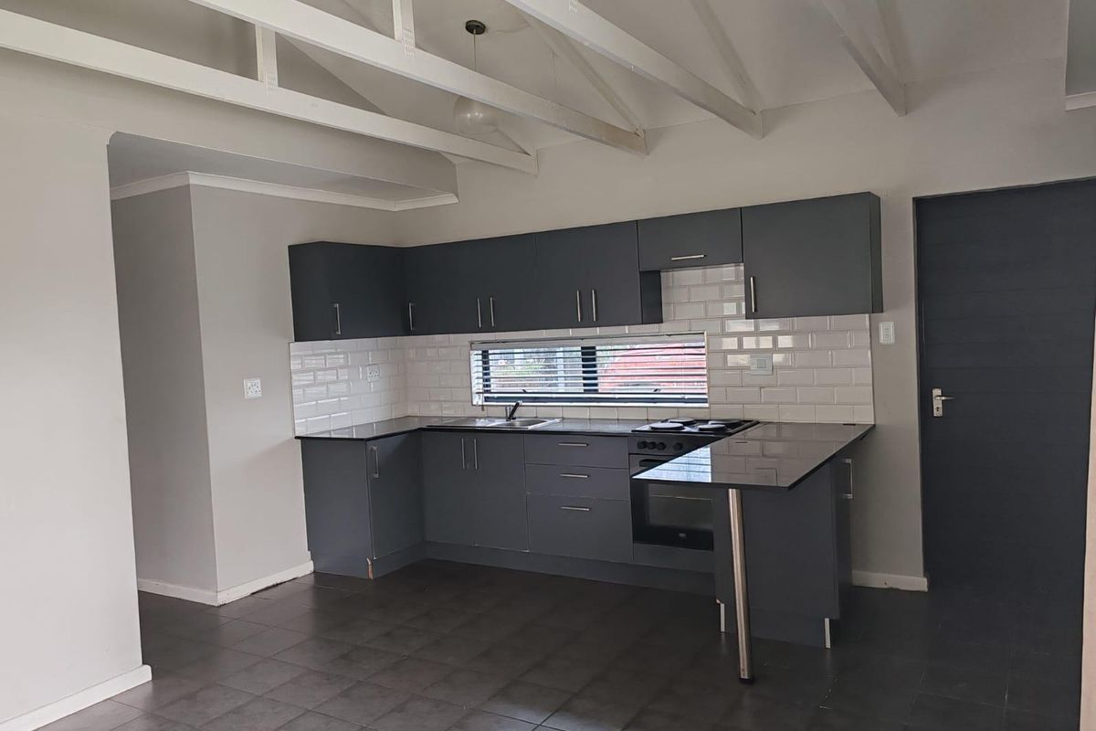 Modern kitchen featuring dark grey cabinetry, subway tile backsplash, stone countertops, built-in oven, hob, and exposed white ceiling beams in an open-plan layout.