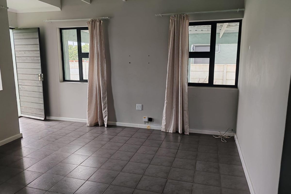 An empty living room featuring tiled flooring, exposed ceiling beams, and natural light through aluminium-framed windows.