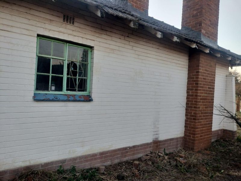 Exterior view of a brick house showing a chimney, painted brick walls, and a damaged window with broken glass and peeling paint.