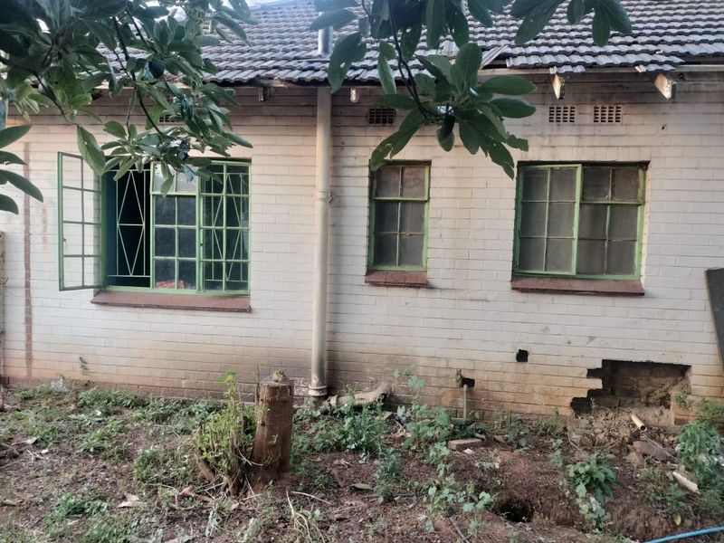 Rear exterior view of a house showing significant structural wear, including damaged brickwork, missing roof tiles, and weathered steel window frames.
