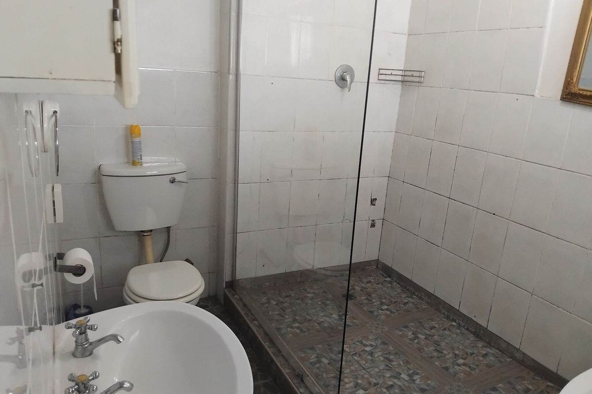 A functional bathroom featuring a glass-enclosed shower, pedestal basin, and toilet, with tiled finishes throughout. The space requires minor maintenance to the shower tiling.