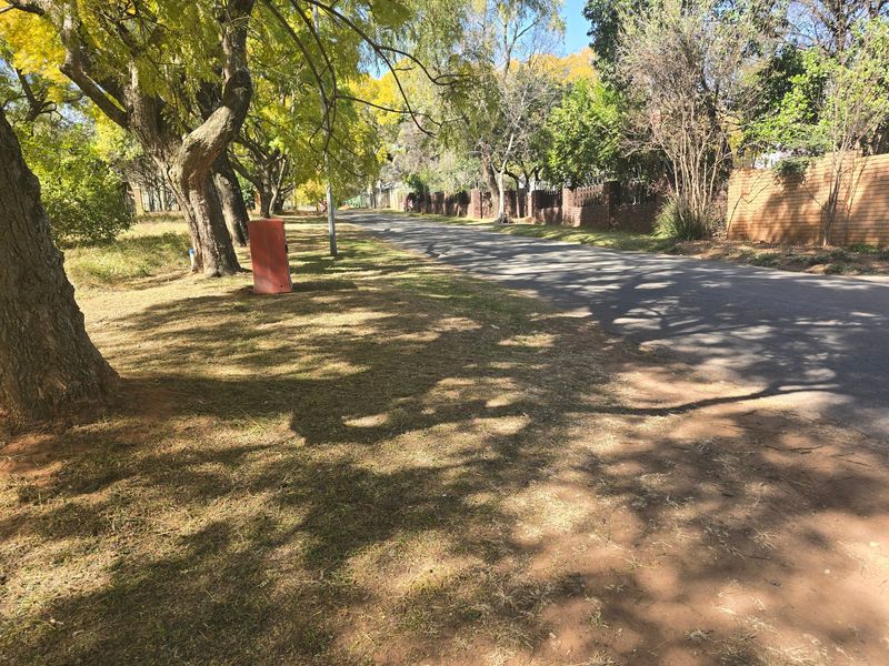A view of the street frontage featuring mature trees, an asphalt road, and a brick boundary wall along the property perimeter.