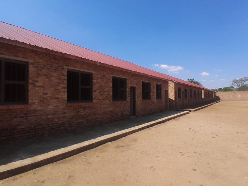 A long, single-story building featuring exposed brick walls, a red corrugated metal roof, and a concrete walkway along the exterior.