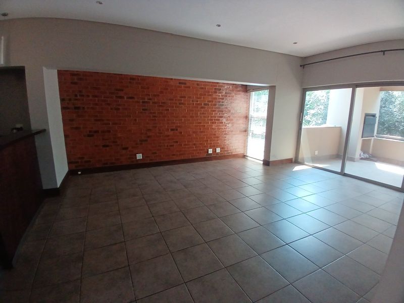 Spacious open-plan living area featuring a striking exposed brick wall, neutral tiled flooring, and large sliding glass doors leading to an outdoor patio.