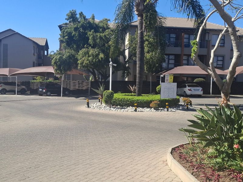 A well-maintained apartment complex exterior featuring paved driveway access, designated shade cloth parking bays, and established landscaped garden areas.