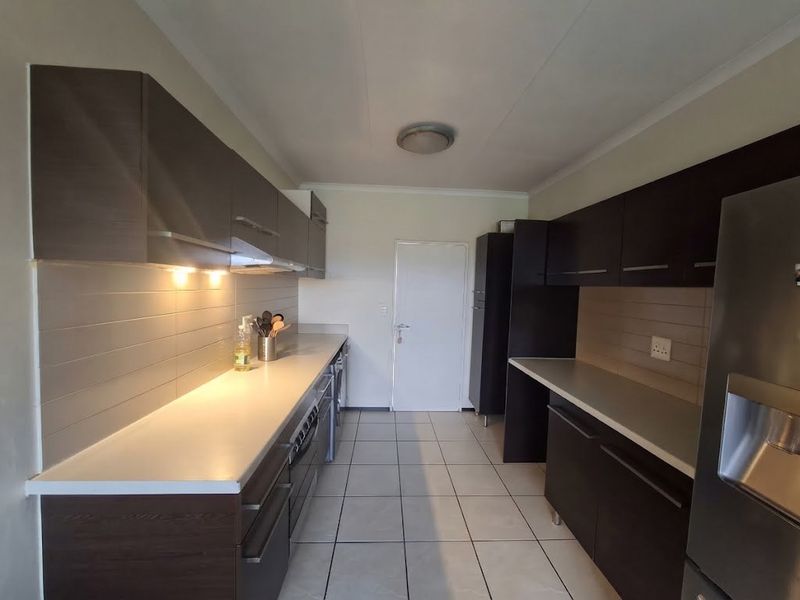 A functional galley-style kitchen featuring dark wood-finish cabinetry, light-colored countertops, tiled backsplash, and ceramic floor tiles with integrated lighting.