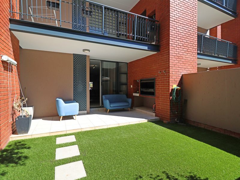 A private rear courtyard featuring a tiled patio, built-in braai, and artificial lawn, with direct access to the interior via sliding glass doors.