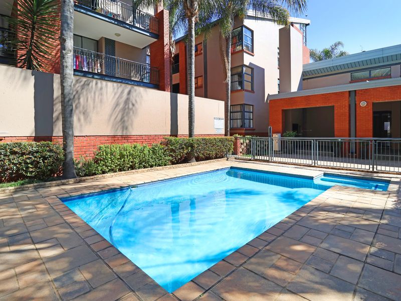 A well-maintained communal swimming pool area featuring paved decking, metal safety fencing, and established palm trees within a townhouse complex.