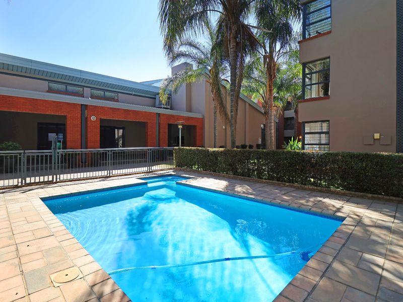 A well-maintained communal swimming pool area featuring paved decking, metal safety fencing, and established garden hedging within a townhouse complex.
