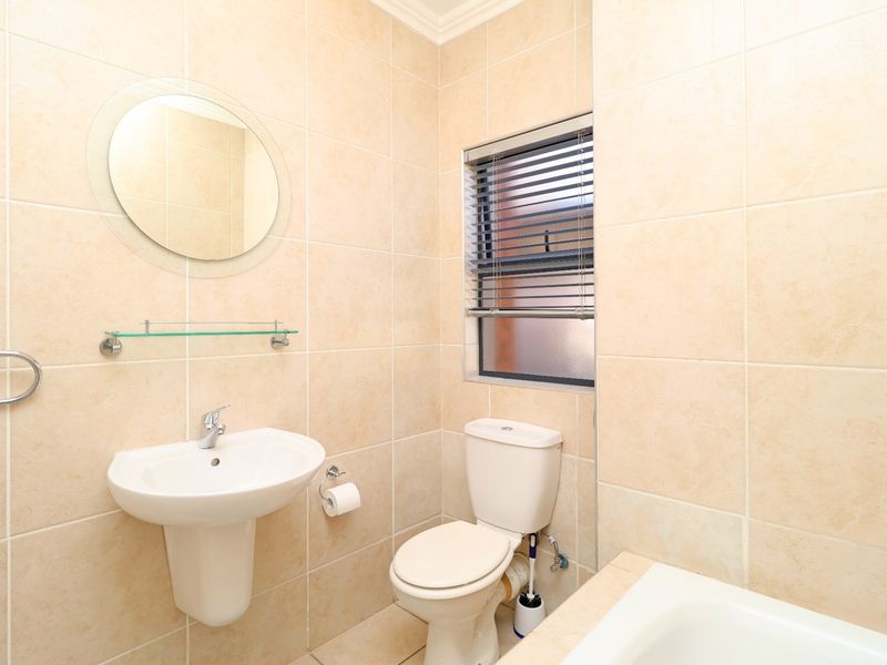 A clean, neutral-toned bathroom featuring a pedestal washbasin, toilet, and bathtub with full-height wall tiling and a window for natural light.