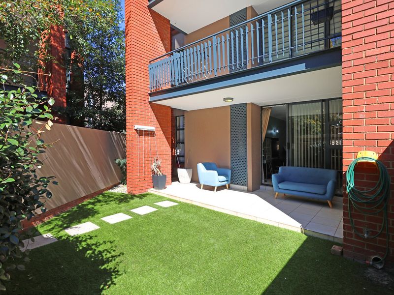 A private rear courtyard featuring a tiled patio area, artificial lawn, and a balcony above with a metal balustrade and brick exterior finish.
