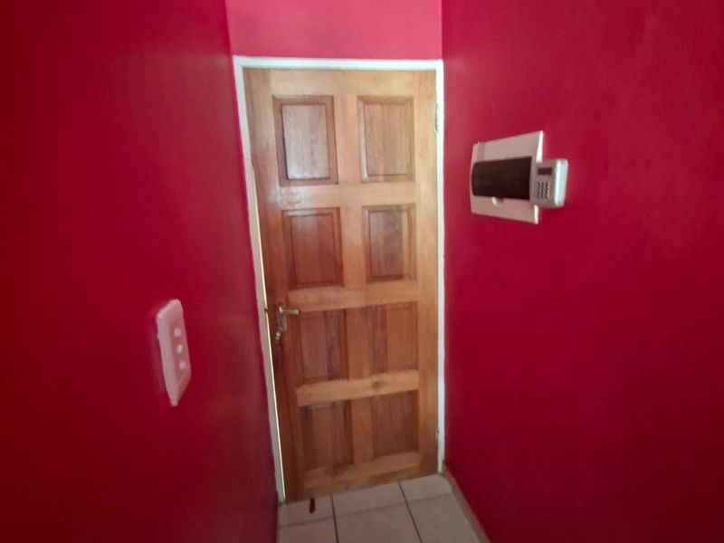 A narrow hallway featuring tiled flooring, a wooden panel door, and a wall-mounted security alarm keypad.