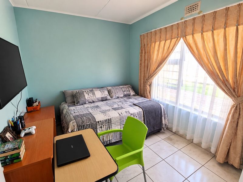 A functional bedroom featuring tiled flooring, a large window with curtains, and a ventilation grille, providing a comfortable living space.