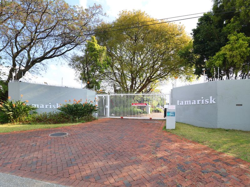 The secure gated entrance to the Tamarisk complex, featuring a brick-paved driveway, perimeter walls with electric fencing, and mature trees.